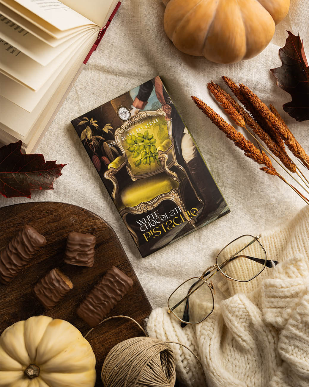 Laurence Tafelschokolade State of the Art weiße Schokolade Pistazie, im Hintergrund einige Riegel, ein Buch, nett angerichtet mit Trockenblumen und einer Tischdecke Laurence Tafelschokolade State of the Art weiße Schokolade Pistazie, im Hintergrund einige Riegel, ein Buch, nett angerichtet mit Trockenblumen und einer Tischdecke