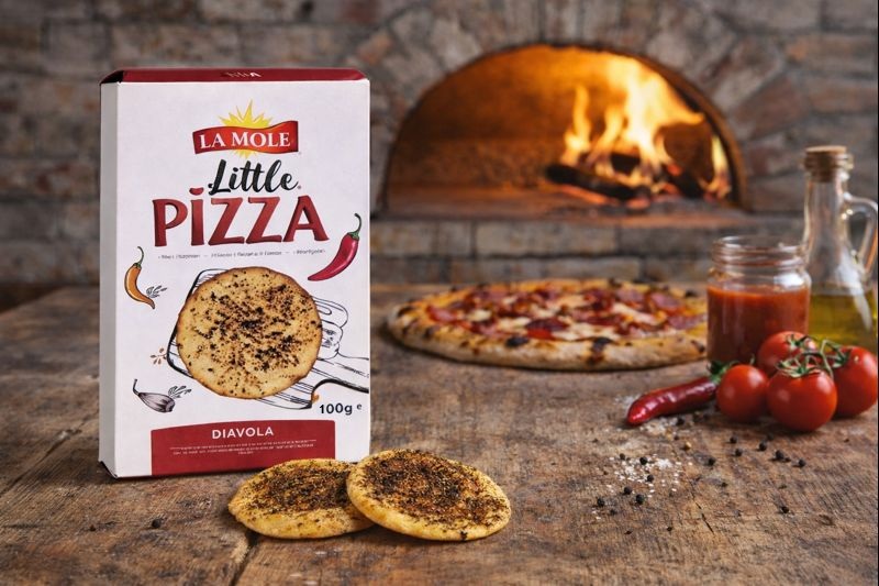 La Mole Little Pizza Diavola Packung mit Inhalt auf einem rustikalen Tisch mit einer frischen Pizza, Olivenöl, Chili-Schote, Tomatensoße und Tomaten, im Hintergrund ein Holzofen La Mole Little Pizza Diavola Packung mit Inhalt auf einem rustikalen Tisch mit einer frischen Pizza, Olivenöl, Chili-Schote, Tomatensoße und Tomaten, im Hintergrund ein Holzofen
