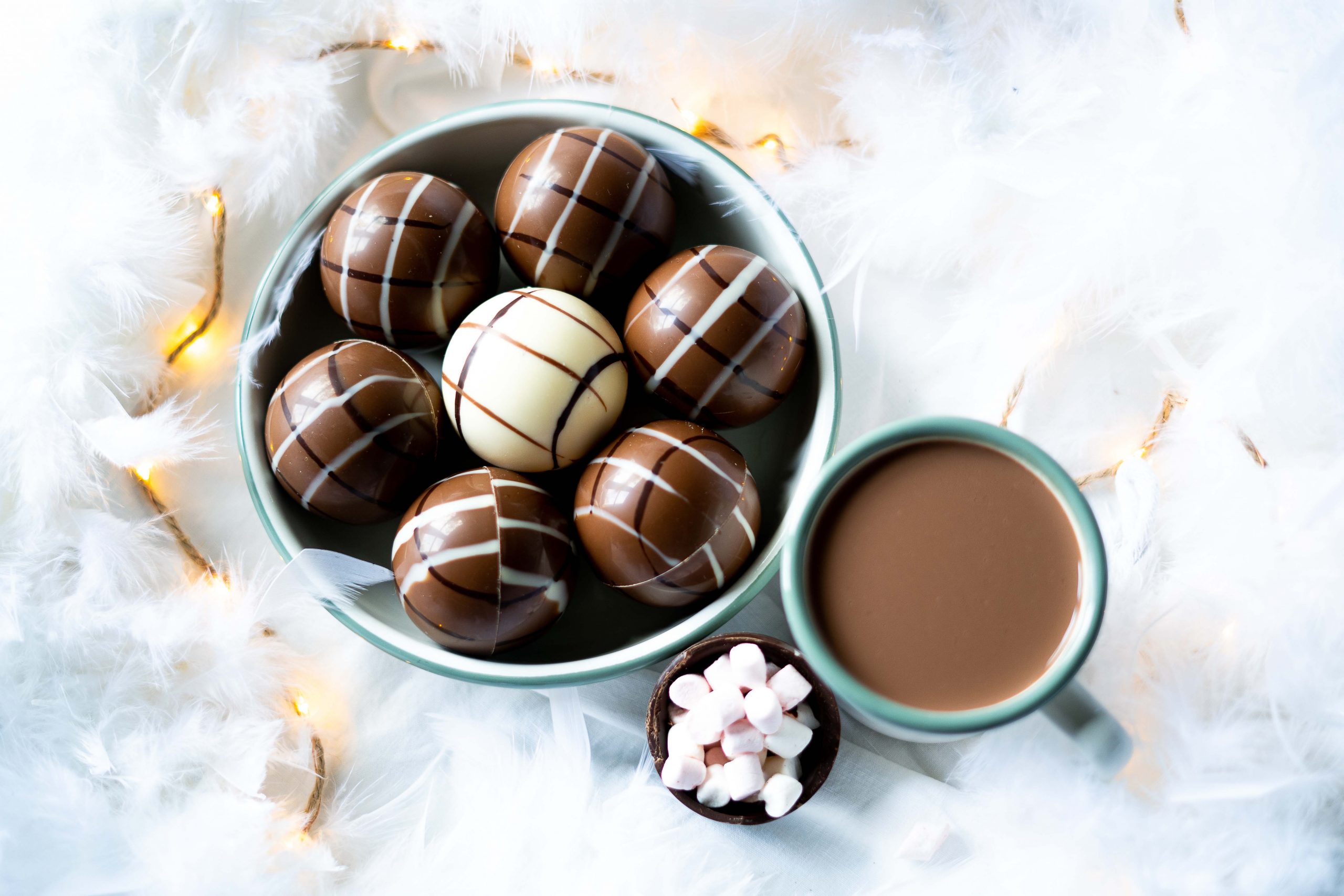 P&T Magic Hot Chocolate Balls in einer Schale, welche auf einem Untergrund mit Federn steht, ein halbierter Chocolate Ball mit Marshmallows, eine Tasse heiße Schokolade P&T Magic Hot Chocolate Balls in einer Schale, welche auf einem Untergrund mit Federn steht, ein halbierter Chocolate Ball mit Marshmallows, eine Tasse heiße Schokolade