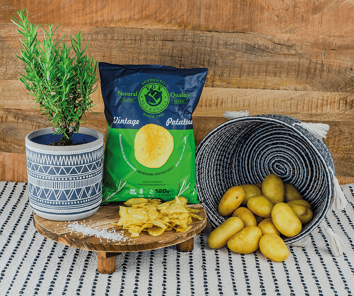 Fox Chips Rosmarin Packung auf einem Holzteller, daneben eine Rosmarin-Pflanze und ein Korb mit Kartoffeln, davor Salzkörner und Chips Fox Chips Rosmarin Packung auf einem Holzteller, daneben eine Rosmarin-Pflanze und ein Korb mit Kartoffeln, davor Salzkörner und Chips