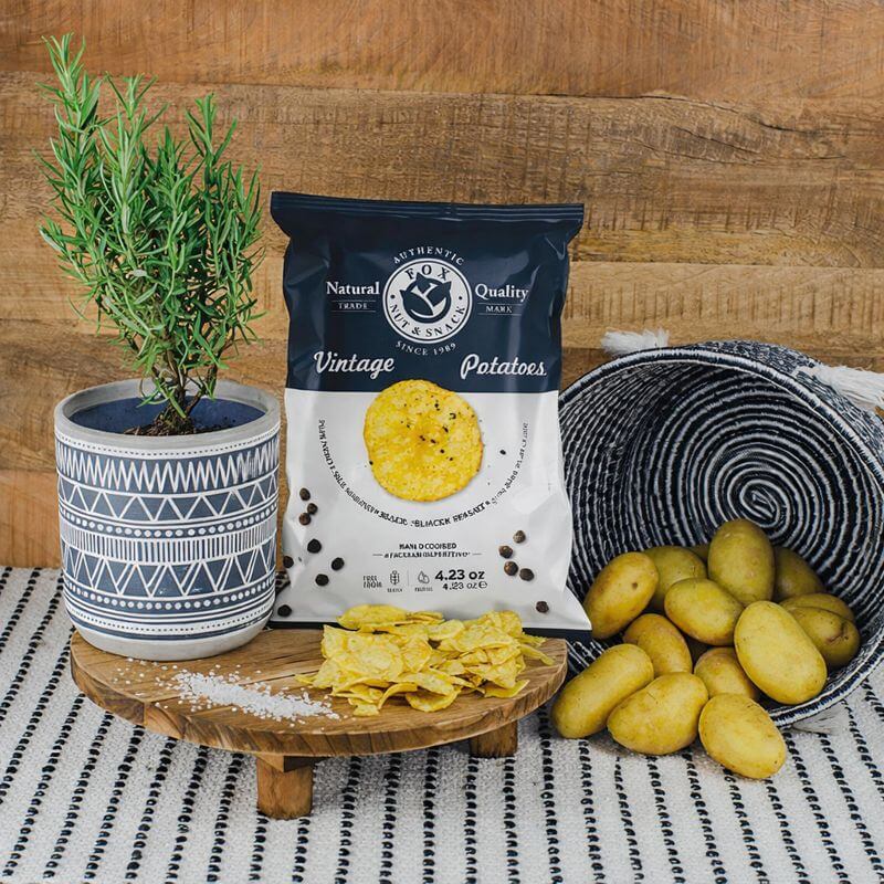 Fox Chips Salz & Pfeffer Packung auf einem Holzteller, daneben eine Pflanze und ein Korb mit Kartoffeln, davor Salzkörner und Chips Fox Chips Salz & Pfeffer Packung auf einem Holzteller, daneben eine Pflanze und ein Korb mit Kartoffeln, davor Salzkörner und Chips