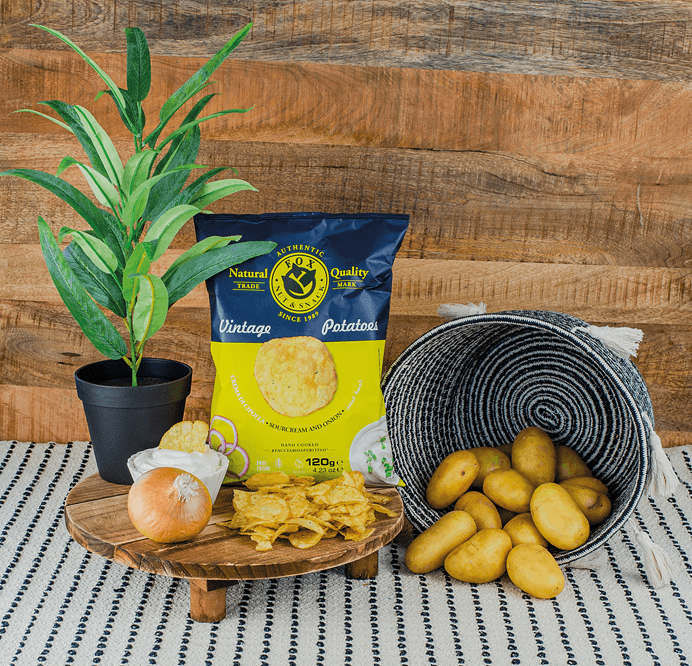 Fox Chips Sourcream & Zwiebel Packung auf einem Holzteller, daneben eine Pflanze und ein Korb mit Kartoffeln, davor eine Zwiebel, eine Schale mit Sourcream-Dip und Chips Fox Chips Sourcream & Zwiebel Packung auf einem Holzteller, daneben eine Pflanze und ein Korb mit Kartoffeln, davor eine Zwiebel, eine Schale mit Sourcream-Dip und Chips