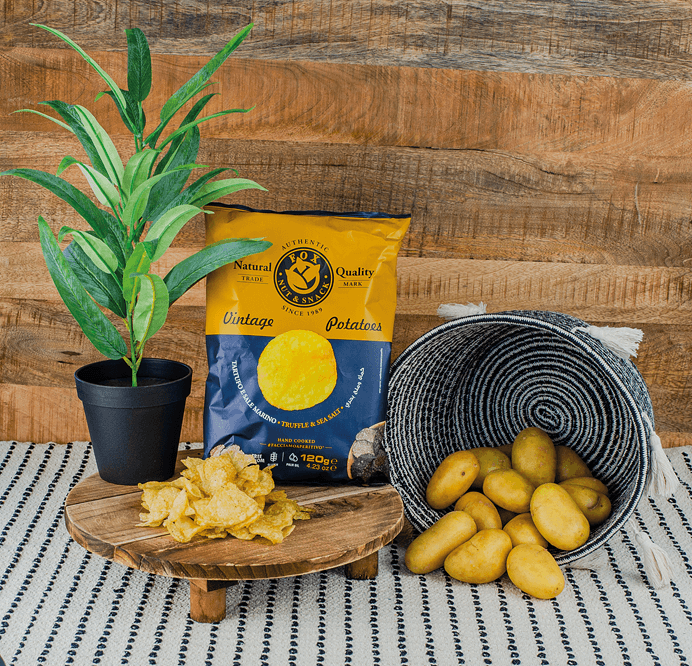 Fox Chips Trüffel Packung auf einem Holzteller, daneben eine Pflanze und ein Korb mit Kartoffeln, davor Chips Fox Chips Trüffel Packung auf einem Holzteller, daneben eine Pflanze und ein Korb mit Kartoffeln, davor Chips
