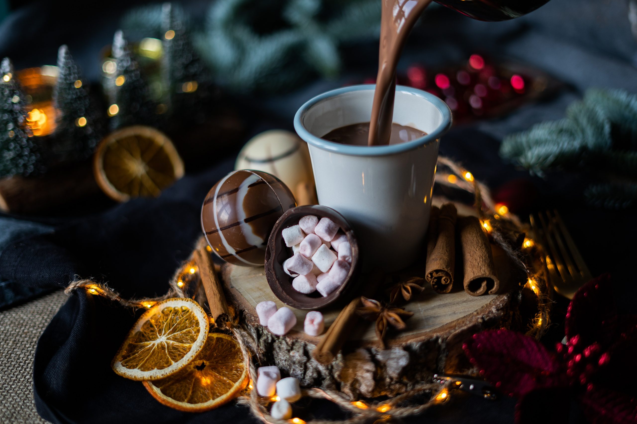 P&T Magic Hot Chocolate Ball und halbierte Variante mit Marshmallows, winterlich angerichtet auf einer Baumscheibe, mit Tannenzweigen, getrockneten Orangenscheiben, Zimtstangen und einem Tischläufer, sowie einer Tasse heißer Schokolade P&T Magic Hot Chocolate Ball und halbierte Variante mit Marshmallows, winterlich angerichtet auf einer Baumscheibe, mit Tannenzweigen, getrockneten Orangenscheiben, Zimtstangen und einem Tischläufer, sowie einer Tasse heißer Schokolade