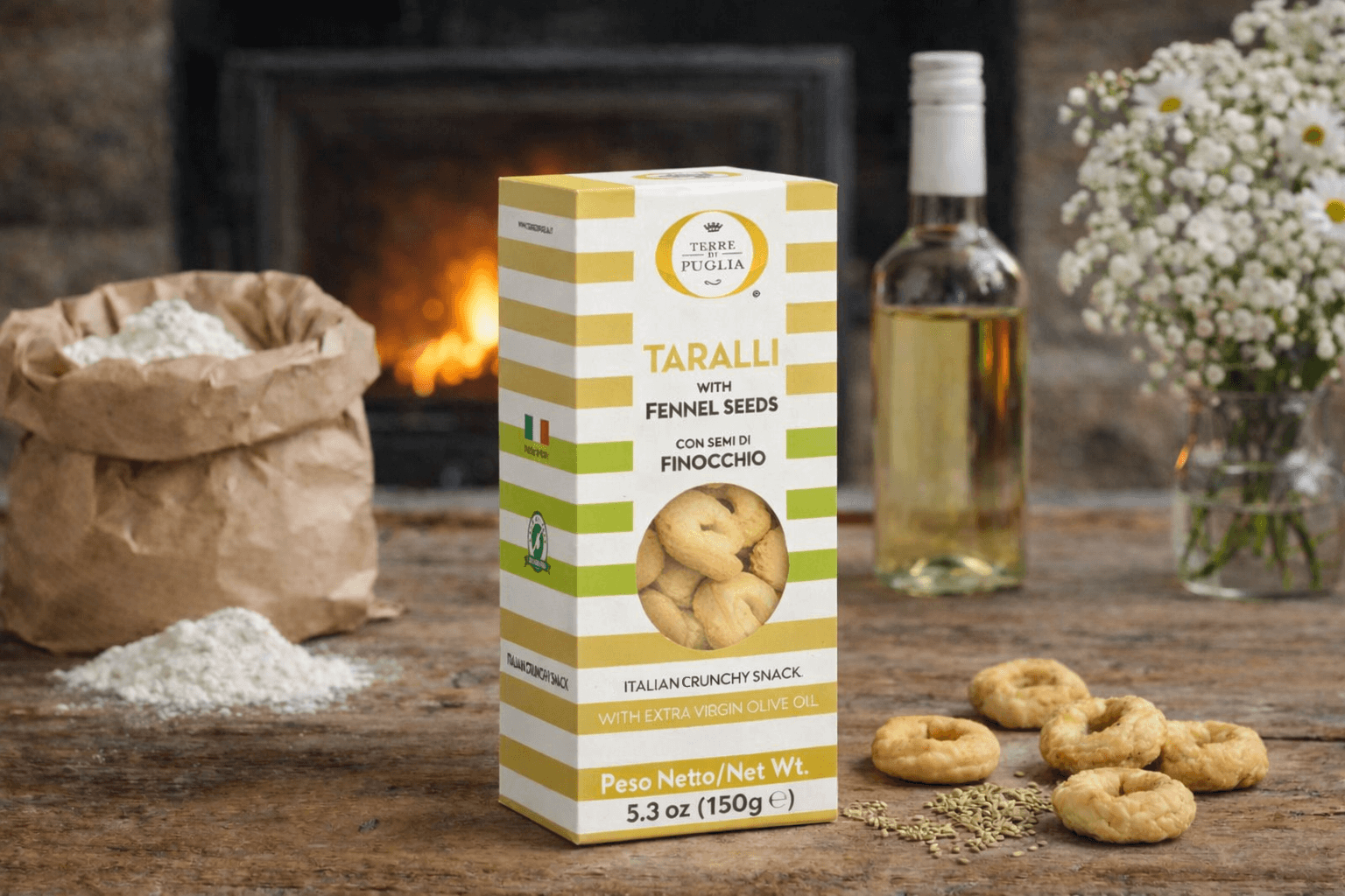 Terre di Puglia Taralli Fenchel Packung, auf einem rustikalen Holztisch, links daneben eine Papiertüte mit Mehl, rechts daneben Taralli-Snacks, Fenchelsamen, eine Flasche Weißwein, eine Vase mit Blumen und im Hintergrund ein Kaminfeuer Terre di Puglia Taralli Fenchel Packung, auf einem rustikalen Holztisch, links daneben eine Papiertüte mit Mehl, rechts daneben Taralli-Snacks, Fenchelsamen, eine Flasche Weißwein, eine Vase mit Blumen und im Hintergrund ein Kaminfeuer