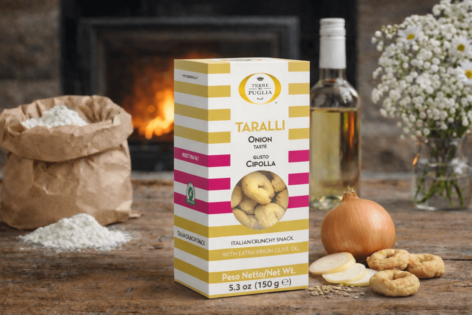 Terre di Puglia Taralli Zwiebel Packung, auf einem rustikalen Holztisch, links daneben eine Papiertüte mit Mehl, rechts daneben Taralli-Snacks, Zwiebeln, eine Flasche Weißwein, eine Vase mit Blumen und im Hintergrund ein Kaminfeuer Terre di Puglia Taralli Zwiebel Packung, auf einem rustikalen Holztisch, links daneben eine Papiertüte mit Mehl, rechts daneben Taralli-Snacks, Zwiebeln, eine Flasche Weißwein, eine Vase mit Blumen und im Hintergrund ein Kaminfeuer